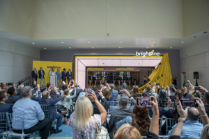 Enthusiastic crowd gathers inside the station, witnessing the exciting moment as the owners cut the ribbon on the impressive Brightline Station entry sign. The atmosphere is filled with anticipation and celebration, marking a significant milestone for the station's grand opening.