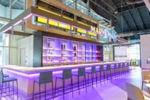 Mary Mary Bar with purple ambient lighting under the bar, in front of the bar stools, inside the Brightline Orlando Train Station, designed by Bigtime Design Studios