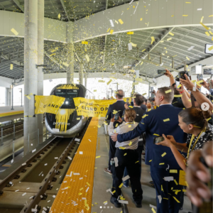 Exciting moment as a Brightline Train triumphantly bursts through a vibrant grand opening ribbon at the station, surrounded by jubilant spectators cheering on the sidelines.