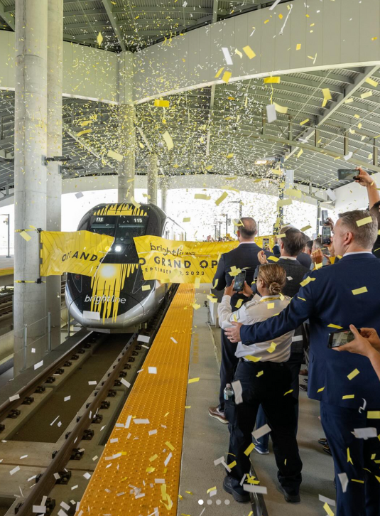 Exciting moment as a Brightline Train triumphantly bursts through a vibrant grand opening ribbon at the station, surrounded by jubilant spectators cheering on the sidelines