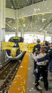 Exciting moment as a Brightline Train triumphantly bursts through a vibrant grand opening ribbon at the station, surrounded by jubilant spectators cheering on the sidelines